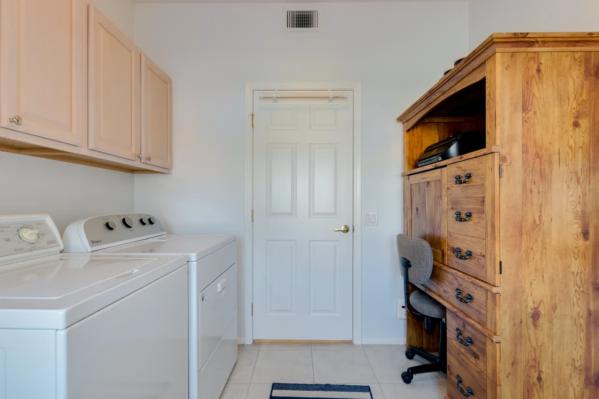 This image presents a practical, multifunctional interior space featuring both a laundry area—with a washing machine and dryer—and an adjacent workspace with an office chair and wooden cabinet. The room feels calm and organized, highlighting the dual-purpose design ideal for extended stays and streamlined routines.