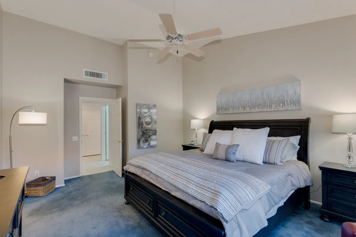 This image features a king-sized bed flanked by nightstands with elegant lamps, decorative wall art, and a ceiling fan overhead. The room is bathed in natural light and exudes a serene, comfortable atmosphere, perfect for rest and privacy.