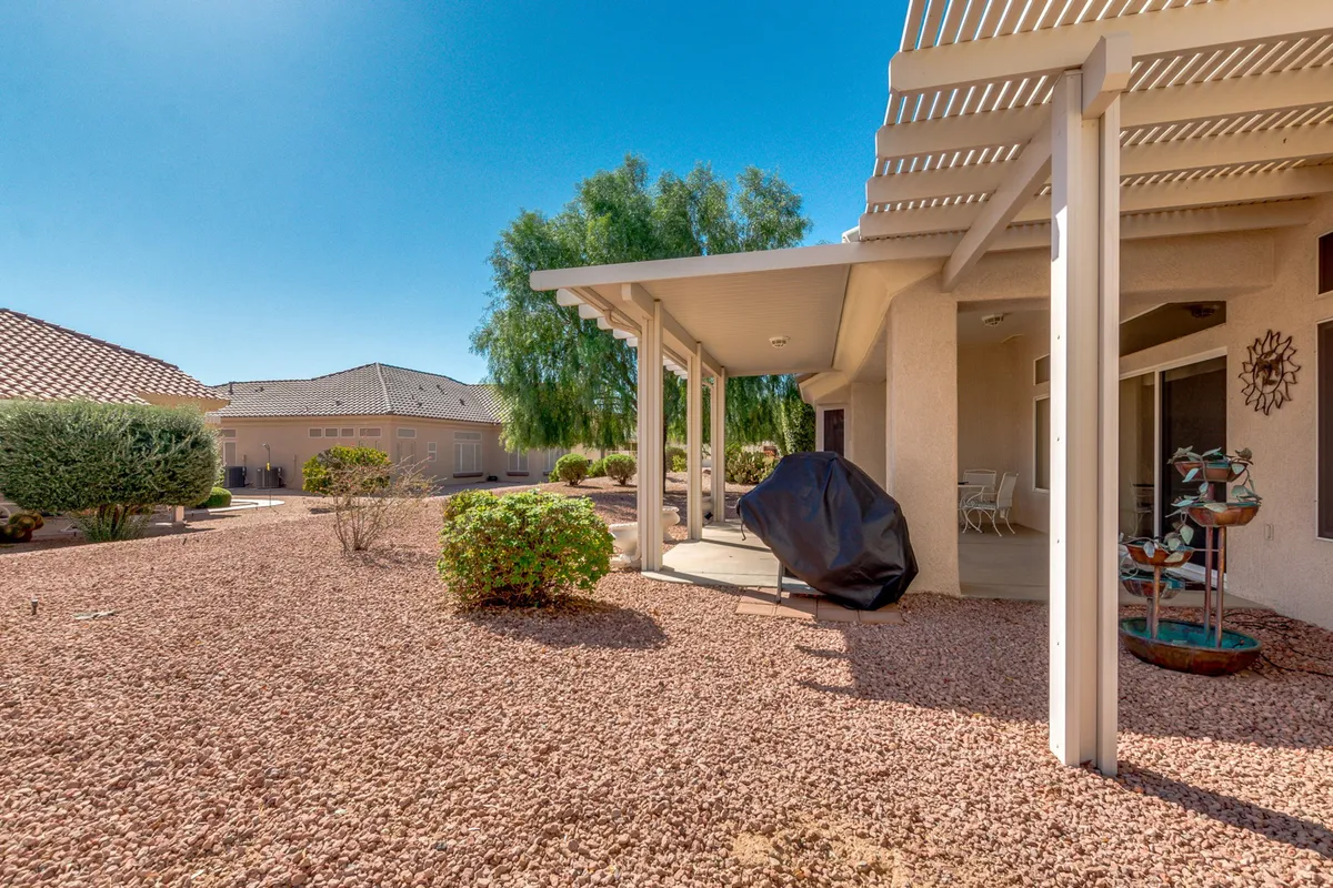 A sunlit backyard featuring a covered patio with a comfortable seating area, a built-in grill for barbecues, a tranquil garden fountain, and lush landscaping. The arrangement invites gatherings for outdoor dining, morning coffee, or relaxing in the shade while surrounded by greenery. The space is oriented to capture abundant sunlight, reflecting the south-facing aspect.