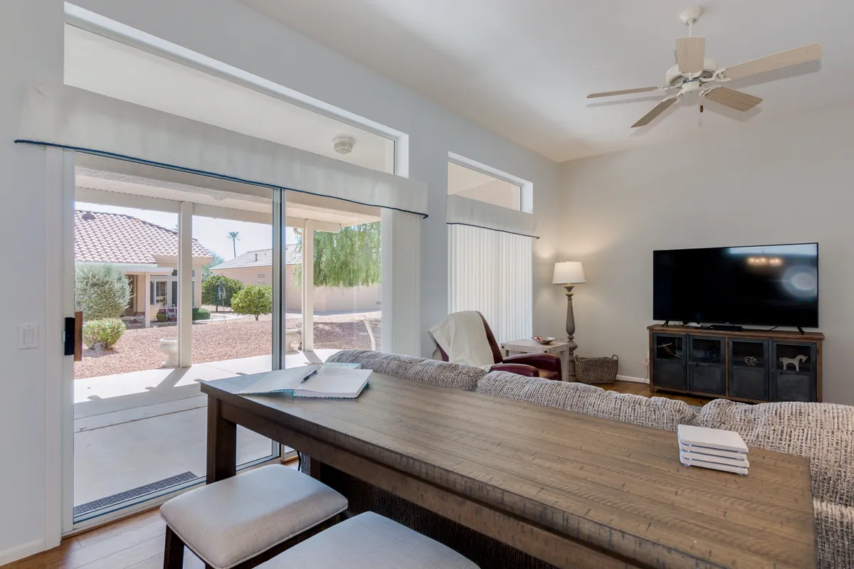 A sunlit dining area with a large table, window views, TV unit, and ceiling fan. The space is set for family meals and gatherings, with plenty of natural light creating a welcoming ambiance.